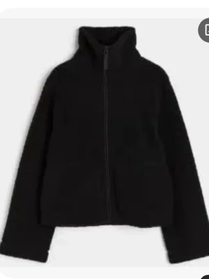 Svart teddyjacka med hög krage - Mysig svart teddyjacka med dragkedja framtill och hög krage. Jackan har två fickor på sidorna och är tillverkad i fluffigt teddy-material som håller dig varm. Perfekt för kyliga dagar och enkel att matcha med olika outfits.