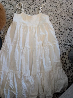 Vit långklänning med smock och tunna band stl 32 - Luftig vit långklänning med smock upptill och tunna axelband som knyts. Klänningen har flera volanger och är tillverkad i ett mjukt, lätt tyg som passar perfekt till varma dagar. Enkel och stilren design utan mönster.