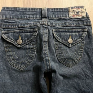 True Religion flared vintage low waist flared jeans - Säljer ett par blå flared vintage jeans från True Religion med ikoniska. Flared och low waisted, has two holes but they are part of the hall vintage vibe. Small Hole in both trouser legs. XS-S size.