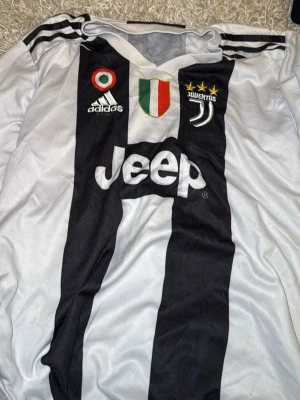 Juventus matchtröja Ronaldo 7 Adidas - Juventus officiella matchtröja från Adidas med Ronaldo 7-tryck på ryggen. Tröjan är vit med svarta ränder, Juventus klubbmärke och sponsorloggor på bröstet. Tillverkad i lätt polyester, perfekt för fotbollsträning eller att bära som supporter.