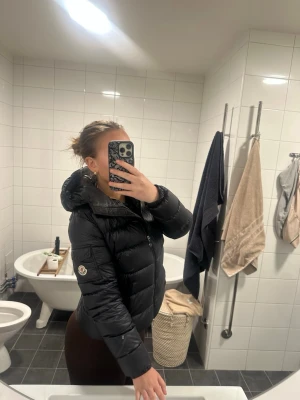 Svart pufferjacka från Moncler - Säljer en svart pufferjacka från Moncler med huva och dragkedja framtill. Jackan har två fickor med dragkedja och den klassiska Moncler-loggan på ärmen. Perfekt för kalla dagar och riktigt snygg till alla outfits. Passar S/M