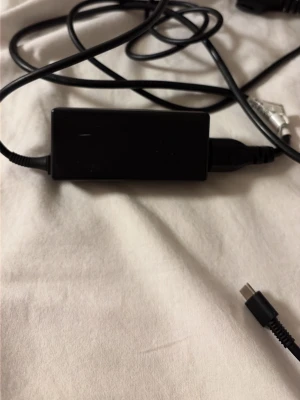 Svart USB-C laddare - En svart USB-C laddare med rektangulär adapter och lång kabel. Perfekt för att ladda laptops, surfplattor eller andra enheter med USB-C port. Sladden är flexibel och adaptern är gjord i plast.