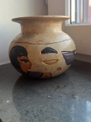 Dekorativ egyptisk vas i keramik - Unik rund vas i beige keramik med handmålade motiv inspirerade av forntida Egypten. Vasen har bred öppning och detaljer som ansikten, symboler och vingar i svart, lila och brunt. Perfekt som inredningsdetalj för dig som gillar antik stil och historia.