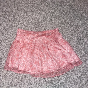 Rosa kortkjol med paisleymönster - Supersöt rosa kortkjol med vitt paisleymönster och lätt volang. Kjolen har en luftig känsla och är perfekt för varma dagar. Resår i midjan för skön passform. Passar dig som gillar en feminin och trendig stil.