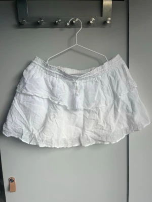 Vit kortkjol med volang och knyt - Söt vit kortkjol i luftig bomull med volangdetaljer och knytband med tofs framtill. Kjolen har resår i midjan för skön passform och en lätt, somrig känsla. Perfekt för dig som gillar en avslappnad och trendig stil.