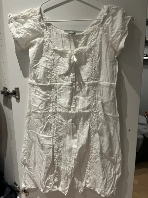Vit kort klänning med spetsdetaljer - Söt vit kortklänning med spetsdetaljer och knyt framtill. Klänningen har korta puffärmar, dekorativa knappar och fina broderade detaljer längs hela framsidan. Perfekt för varma dagar och har en romantisk vibe. Har haft denna flera gånger på skolavslutning och student, så den passar till många tillfällen. Med klänningen kommer en underklänning då själva klänningen är lite genomskinlig. Jag tycker den är perfekt längd för mig som är ca 170, kort men inte för kort. Sen finns det två fläckar,maskros