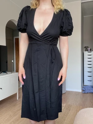 Svart omlottklänning med puffärm från Cubus, storlek XS (nyskick) - Svart omlottklänning med djup v-ringning och puffiga kortärmar. Knytband i midjan och går ner till knäna. Använd en gång, superfint skick!