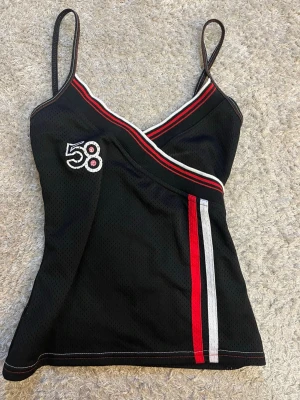 Svart sportig linnetopp med siffran 58 - Snygg svart linnetopp med tunna axelband och omlott-ringning. Dekorerad med siffran 58 på bröstet och röda samt vita ränder längs ringningen och framtill. Tillverkad i ett mesh-liknande syntetmaterial som ger en sportig vibe.
