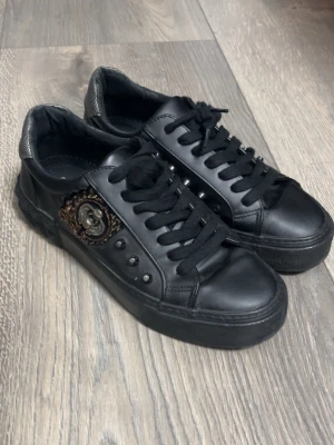 Svarta sneakers med emblem och nitar - Säljer ett par svarta sneakers i läder-look med coolt metall-emblem och dekorativa nitar på sidan. Skorna har svarta skosnören och en chunky sula. Perfekta för dig som gillar detaljer och vill sticka ut lite extra.