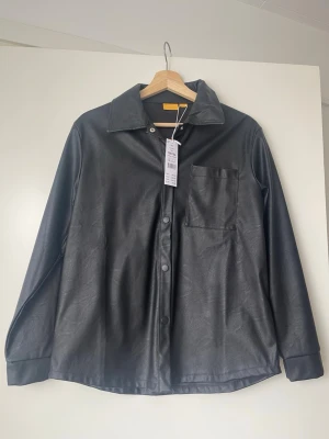 Svart overshirt i PU-läder - Stilren svart overshirt från gina tricot i mjukt PU-läder. Skjortan har klassisk krage, tryckknappar framtill och en bröstficka. Perfekt för dig som vill ha en trendig och clean look med en touch av edge.