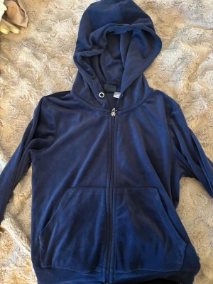 Mörkblå hoodie från Gina Tricot - Mysig mörkblå hoodie från Gina Tricot i mjukt velourmaterial. Dragkedja framtill, två stora fickor och en rejäl huva med metallöljetter. Perfekt för chill dagar eller när du vill ha något bekvämt och snyggt.
