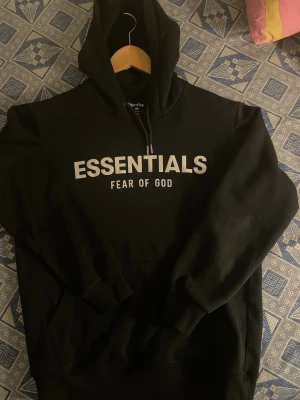 Svart Essentials Fear of God hoodie - Svart hoodie från Essentials Fear of God med stor vit logga på bröstet. Klassisk passform med huva och känguruficka. Tillverkad i mjuk bomull och polyesterblandning, perfekt för en avslappnad och trendig streetwear-look.