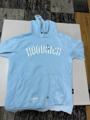 Ljusblå hoodie från Hoodrich - Säljer en ljusblå hoodie från Hoodrich med stor vit logga på bröstet och känguruficka framtill. Hoodien har huva med dragsko och ribbade muddar. Tillverkad i mjukt bomullsmaterial som känns skönt mot huden. Perfekt för en chill och avslappnad stil.