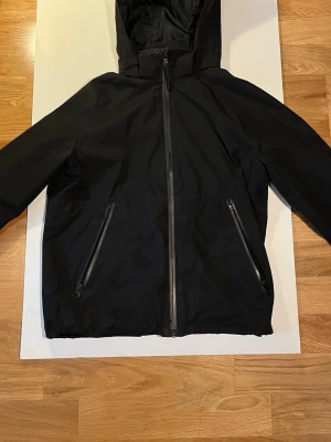 Svart windbreaker med huva - Svart windbreaker med dragkedja hela vägen ner och två fickor med dragkedja. Jackan har en luva.