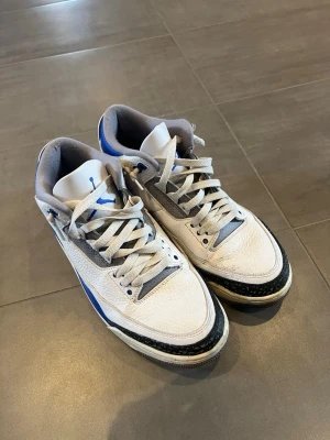Air Jordan 3 Retro White/Blue - Säljer ett par Air Jordan 3 Retro sneakers i vitt med blå och svarta detaljer. Skorna har klassisk siluett med grå mesh och ikoniskt elefantmönster på tån och hälen. Tjock sula och Jumpman-logga på plösen. Perfekta för dig som gillar streetwear och sneakerskultur. Storlek 44, box tillkommer