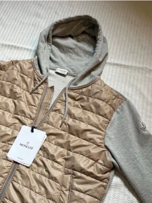 Moncler cardigan beige - Snygg cardiganjacka från Moncler, perfekt för sommarkvällar!  Alla tags och nfs finns, beige quiltad front och rygg i glansigt material samt gråmelerade ärmar och huva i mjuk bomull. Nästan helt ny bara använd ett fåtal gånger! Jackan har dragkedja och fickor fram. Perfekt för lager-på-lager. Pris kan diskuteras!