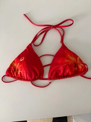Röd blommig triangelbikini - Snygg röd trekantsbikini med stora blommor i orange och gul nyans. Aldrig använd men dock har prislappen klippts bort!
