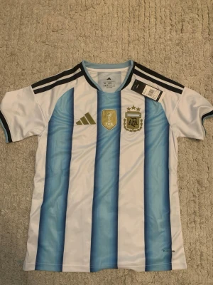 Argentina matchtröja Adidas  - Säljer en officiell Argentina matchtröja från Adidas med klassiska blå och vita ränder, broderat AFA-emblem och tre stjärnor ovanför. Tröjan har svarta axelränder och FIFA-märke på bröstet. Tillverkad i lätt och ventilerande material, perfekt för fotboll.
