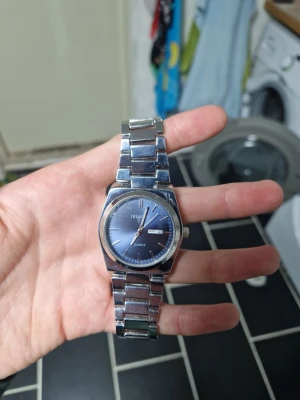 Blå IRLA Quartz klocka i metall - Stilren klocka från IRLA med blå urtavla och dag- och datumvisning. Armbandet är i blankt silverfärgat metall och har en klassisk länkdesign. Perfekt accessoar för dig som gillar en clean look. Kommer med justerare