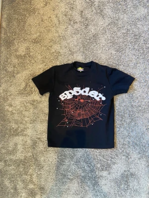 Svart Sp5der Hoodie – Stilren och iögonfallande - Svart Sp5der hoodie med den klassiska SP5DER-loggan i vit text på framsidan. Hoodien har ett unikt designmönster med röda spindelnät och röda stjärnor som skapar en exklusiv och modern streetwear-look. Tillverkad i ett mjukt och bekvämt material som passar perfekt för både vardag och fritid. Ett snyggt plagg för dig som vill sticka ut med stil och samtidigt ha hög komfort