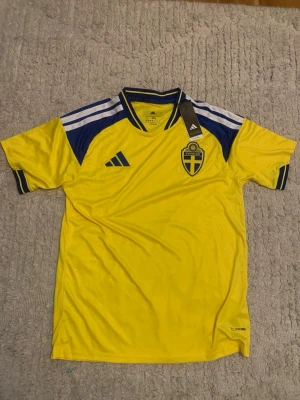 Sverige matchtröja Adidas gul/blå - Säljer en officiell Sverige matchtröja från Adidas i gult och blått med Svenska Fotbollförbundets emblem på bröstet. Tröjan har ClimaCool-material som andas, korta ärmar och klassiska Adidas-ränder på axlarna. Perfekt för fotbollsfans!