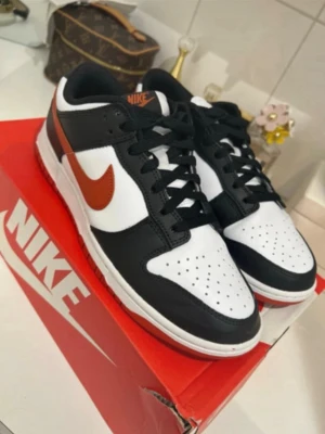 Nike Dunk Low svart/vit/röd - Nike Dunk Low sneakers i svart och vitt läder med röda detaljer och klassisk Swoosh på sidan. Skorna har perforerad tåbox, svart snörning och vit mellansula. Perfekt för dig som gillar streetwear och vill ha en ikonisk look.