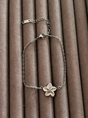 Silverfärgat armband med blomma - Ett tunt och stilrent armband i silverfärgad metall med en detaljrik blomma i mitten. Armbandet har en justerbar kedja och ett klassiskt klolås. Perfekt för dig som gillar diskreta men snygga accessoarer.