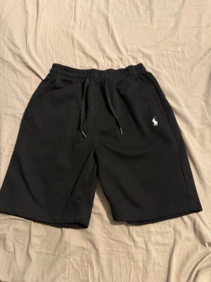Svarta shorts från Polo Ralph Lauren - Svarta shorts från Polo Ralph Lauren med klassisk logga broderad i vitt på vänster ben. Shortsen har elastisk midja med snörning och sidofickor. Perfekta för en avslappnad och stilren look. QR koden fungerar