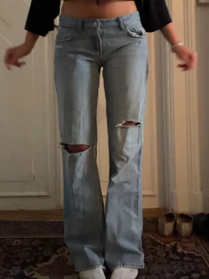 Lågmidjade jeans - Säljer ett par ljusblå  jeans med slitningar och hål vid knäna. Jeansen har klassisk femficksmodell, hög midja och raka ben som går ut lite nedtill. Perfekta för en avslappnad och trendig look.