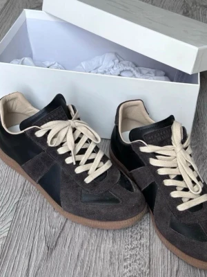 Maison Marigela Gats Bruna - Snygga sneakers i svart läder och mörkgrå mocka med beige snörning och bruna gummisulor. Klassisk siluett med retrovibbar och kontrasterande materialval. Perfekta för dig som gillar stilrena och tidlösa sneakers.