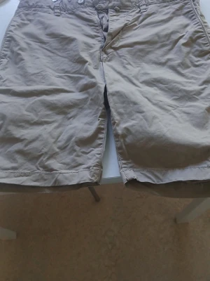 Beige bomullsshorts med fickor - Snygga beige shorts i bomull med klassisk passform. De har framfickor, bakficka med knapp och bälteshällor. Perfekta för varma dagar och enkel att matcha med olika stilar.