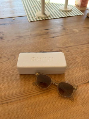 Beiga solglasögon från CHIMI - Snygga solglasögon från CHIMI med transparent beige båge och fyrkantig form. Glasen är mörka och ger en cool look. Kommer med ett stilrent vitt fodral med CHIMI-logga. Perfekta för att höja din outfit till nästa nivå.