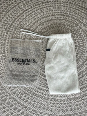 Grå Essentials Fear of God mjukisbyxor - Snygga ljusgrå mjukisbyxor från Fear of God Essentials med tryckt logga på benet. Byxorna har resår i midjan med dragsko, sidofickor och muddar vid bensluten. Perfekta för en chill och trendig look. Alla tags sitter kvar, helt nytt skick och aldrig använda. Anledning är för att det var för korta för mig.