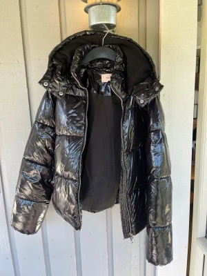 Svart glansig pufferjacka från Nelly - Säljer en svart, glansig pufferjacka från Nelly med stor huva och dragkedja framtill. Jackan har en fin och unik look och breda kanaler som ger en cool och trendig vibe. Perfekt för kalla vinterdagar när du vill hålla stilen. Använd endast en vinter