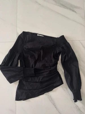 Svart offshoulder blus - Trendig svart offshoulder blus med smockdetaljer vid axlar och ärmslut. Snygg asymmetrisk skärning i midjan och tillverkad i ett glansigt tyg som ger en lyxig känsla. Storlek M men sitter som S!!💖💖