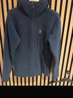 Mörkblå fleecejacka från Haglöfs - Mysig mörkblå fleecejacka från Haglöfs med huva och hel dragkedja. Jackan har två sidofickor med dragkedja och en bröstficka med Haglöfs-logga. Perfekt för kalla vinterdagar och outdoor-äventyr.