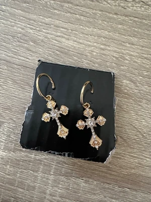 Guldörhängen med kors och stenar - Snygga örhängen i guldfärg med hängande kors dekorerade med små glittrande stenar. Korsen har detaljerad design och örhängena har en enkel krok för att sätta i örat. Perfekta för dig som gillar statement-smycken med lite edge. OBS: FRAKTAR INOM 24H!🩷