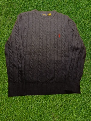 Polo Ralph Lauren stickad tröja - Kabelstickad marinblå tröja från Polo Ralph Lauren med klassisk röd logga broderad på bröstet. Tröjan har rund halsringning, ribbade muddar och nederkant samt diskreta sidoslitsar. Perfekt för dig som gillar tidlös och stilren design. OBS liten defekt vid på bild 5 men inget som syns vid användning.