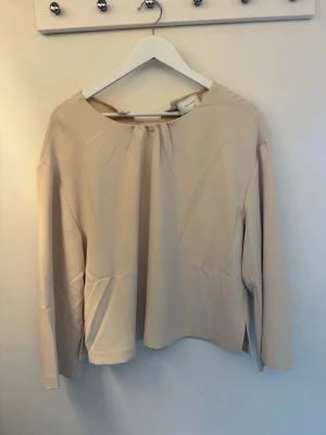 Beige långärmad blus från neo noir - Stilren beige blus från neo noir med rundad halsringning och diskret rynk vid halsen. Blusen har långa ärmar och en rak, något lös passform. Materialet ser ut att vara mjukt och lätt, perfekt för en clean och enkel look.