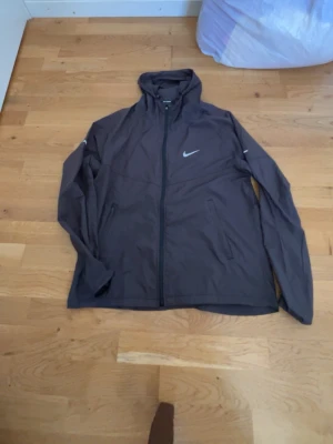Svart vindjacka från Nike med huva - Svart vindjacka från Nike med dragkedja framtill, huva och två sidofickor med dragkedja. Jackan har Nike-logga på bröstet och är tillverkad i ett lätt, vindtätt material. Perfekt för blåsiga dagar och sportiga outfits.