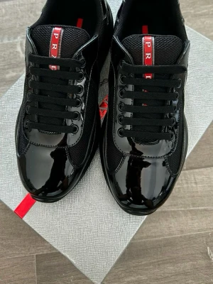 Svarta sneakers från Prada - Snygga svarta sneakers från Prada med glansiga lackdetaljer och meshpaneler. Skorna har svarta skosnören och en röd logotyp på plösen. Perfekt för dig som vill ha en stilren och exklusiv look med streetkänsla.