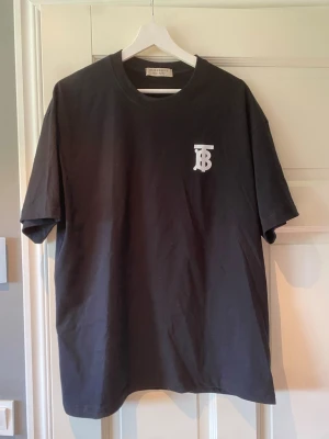Svart Burberry t-shirt - Snygg svart t-shirt från Burberry med klassisk vit TB-logga på bröstet. T-shirten har rund halsringning och lite oversize passform. Tillverkad i mjuk bomull som känns skön mot huden. Perfekt för en clean och stilren look.
