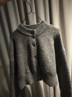 Grå kofta från ZARA med knappar - Mysig grå kofta från ZARA med rund halsringning och stora knappar framtill. Koftan är stickad i ett fluffigt material och har långa ärmar samt ribbade detaljer vid hals och ärmslut. Perfekt för lager-på-lager och enkel att styla.
