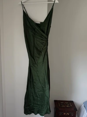 Grön siden omlottklänning från Zara - Mörkgrön långklänning från Zara med omlottdesign och tunna axelband. Klänningen har en elegant v-ringning och är tillverkad i ett glansigt material som ger en lyxig känsla. Perfekt för dig som vill ha en stilren och trendig look.