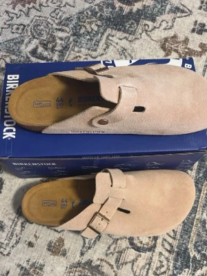 Birkenstock Boston sandfärgade tofflor - Säljer ett par Birkenstock Boston tofflor i sandfärgad mocka. Klassisk slip-in modell med justerbart spänne och mjuk innersula i kork. Perfekta för dig som vill ha både stil och komfort. Levereras i originalkartong.