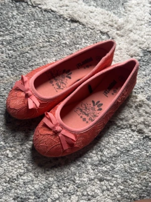 Korallfärgade ballerinaskor med spets - Säljer ett par söta ballerinaskor från Primark i korallrosa med spetsdetaljer och små rosetter framtill. Skorna har rund tå och tunn gummisula. Perfekta för dig som gillar en feminin och lekfull stil.