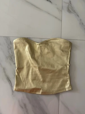 Guldig axelbandslös tubtopp - Snygg guldig tubtopp med bandeau-modell och lätt glansigt material. Toppen är axelbandslös och har en enkel, stilren design som passar perfekt till trendiga outfits. 