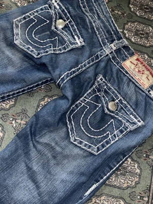 True Religion blå jeans med kontrastsömmar - Säljer ett par blå jeans från True Religion med tydliga vita kontrastsömmar och stora bakfickor med ikonisk brodyr. Jeansen har en klassisk femficksmodell och silverfärgade knappar. Materialet är denim i bomull med tvättad look.
