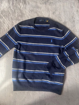 Marinblå randig stickad tröja Polo - Snygg marinblå stickad tröja från Polo Ralph Lauren med tunna vita och blå ränder. Klassisk rund halsringning och broderad logga på bröstet. Perfekt för dig som gillar stilrena och tidlösa plagg.