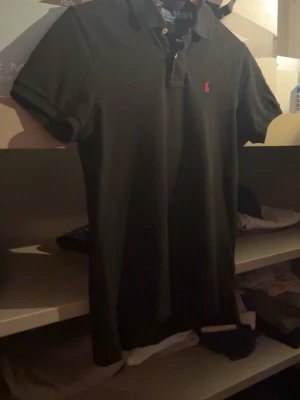 Svart pikétröja från Ralph Lauren - Klassisk svart pikétröja från Ralph Lauren med röd broderad logga på bröstet. Slim fit-modell med korta ärmar och krage. Perfekt till jeans eller shorts för en clean och stilren look.